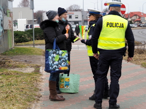 policjantka z policjantem w kamizelkach odblaskowych przekazuje opaski odblaskowe pieszym na chodniku