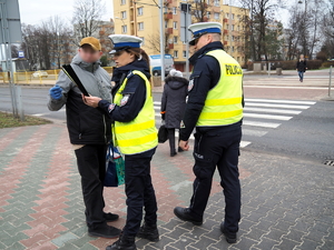 policjantka z policjantem w kamizelkach odblaskowych przekazuje opaski odblaskowe pieszym na chodniku