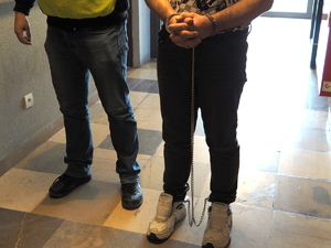 policjant prowadzi po korytarzu pod ramię zatrzymanego, który ma założone kajdanki na ręce i nogi