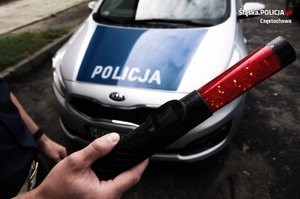 policyjny radiowóz w tle, przed nim zbliżenie na urządzenie do badania stanu trzeźwości, trzymane w ręce przez policjanta