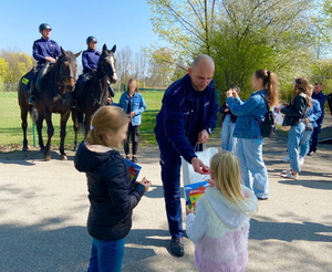 policjant przekazuje książeczki edukacyjne dzieciom, w tle policjanci na koniach pozują do zdjęcia z nastolatką