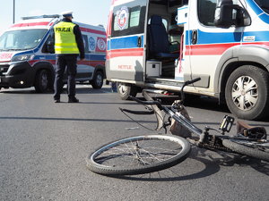 rower leży na jezdni, w tle policjant idzie w kierunku karetki pogotowia, która stoi obok roweru