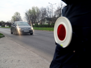 tarcza do zatrzymywania pojazdów wystaje z kieszeni spodni policjanta, w tle droga i poruszający się nią samochód osobowy