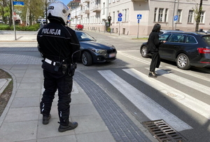 policjant w mundurze i kaskiem na głowie stoi przy przejściu dla pieszych - po pasach przechodzi piesza, samochody osobowe ustepują jej