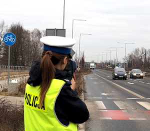 policjantka w kamizelce odblaskowej trzyma w ręce ręczny miernik prędkości i mierzy prędkość samochodów przejeżdżających w rejonie przejścia dla pieszych