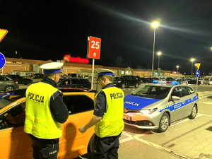 2 policjantów kontroluje pomarańczowy samochód na parkingu przy sklepie