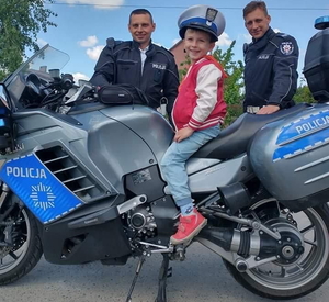 chłopczyk siedzi na motocyklu policyjnym w czapce policyjnej, uśmiecha się. Za nim stoją policjanci z ruchu drogowego
