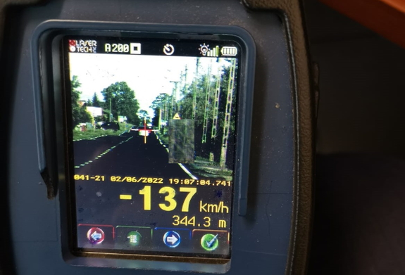 zdjęcie przedstawia przeprowadzanie policyjnym radarem pomiaru prędkości pojazdu BMW oddalającego się. Widoczny jest też wynik pomiaru, który wskazuje prędkość 137 kilometrów na godzinę