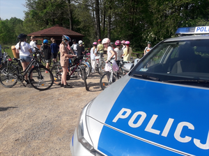 na zdjęciu na pierwszym planie widnieje napis Policja na radiowozie, na drugim planie dzieci na rowerach