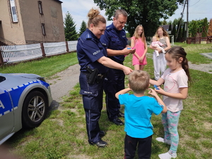 policjanci na podwórku rozdają dzieciom opaski odblaskowe, w tle dorosłe kobiety i radiowóz