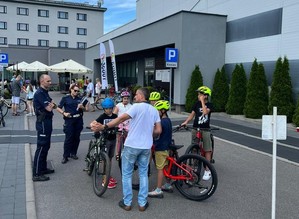 na zdjęciu stoi umundurowany funkcjonariusz i funkcjonariuszka obok na rowerach po prawej stronie stoją młode osoby z rowerami