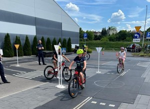 na zdjęciu dzieci na rowerach jeżdżą na miasteczku rowerowym na którym znajduję sią ronda i skrzyżowania. W tle policjanci oraz egzaminator.