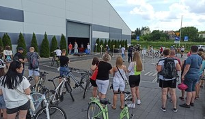 na pierwszym planie zaparkowane rowery oraz dzieci i rodzice czekające na egzamin. na drugim planie grupa dzieci jeżdżąca na rowerach w trakcie trwania egzaminu