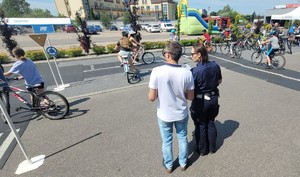 na pierwszym planie policjantka ubrana w granatowy mundur oraz stojący obok egzaminator ubrany w niebieskie spodnie oraz białą koszulkę. Na drugim planie grupa dzieci na rowerze na placu miasteczka rowerowego pokonują skrzyżowanie