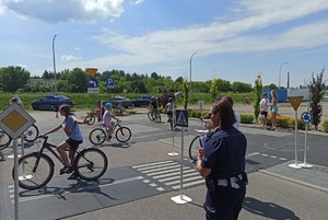 na pierwszym planie policjantka ubrana w granatowy mundur. Na drugim planie grupa dzieci na rowerze na placu miasteczka rowerowego pokonują skrzyżowanie