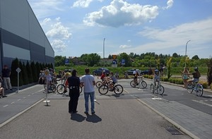 na pierwszym planie policjantka ubrana w granatowy mundur oraz stojący obok egzaminator ubrany w niebieskie spodnie oraz białą koszulkę. Na drugim planie grupa dzieci na rowerze na placu miasteczka rowerowego pokonują skrzyżowanie