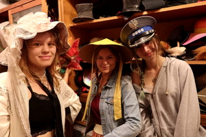 na zdjęciu 3 młode dziewczyny w przebieralni teatru. Każda ma ubrany inne nakrycie głowy. Pierwsza z lewej ma założony czepiec, druga chiński kapelusz a trzecia policyjną czapkę gabardynową.
