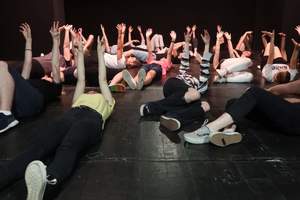 na zdjęciu grupa kilkunastu młodych osób leżąca na plecach na deskach sceny teatralnej, trzymająca obie wyprostowane ręce ku górze