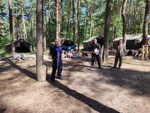 na zdjęciu policjant, strażak oraz leśniczy przeprowadzając prelekcję dla harcerzy, którzy siedzą półkolem na leśnej polanie