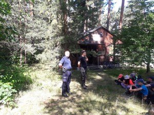 na zdjęciu policjant, strażak oraz leśniczy przeprowadzając prelekcję dla harcerzy, którzy siedzą półkolem na leśnej polanie