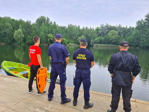 na zdjęciu policjant, strażak, funkcjonariusz straży miejskiej oraz ratownik wodny stoją tyłem do zdjęcia patrząc na wodę