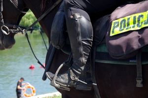 zbliżenie na buty policyjnego jeźdźca, który siedzi na koniu,. w tle zbiornik wodny i napis policja na siodle