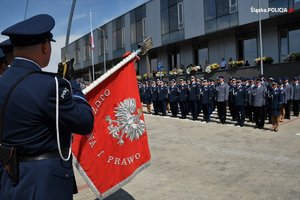 policjant ze sztandarem stoi na placu podczas uroczystości, zbliżenie na sztandar koloru czerwonego z napisem Ojczyzna i Prawo z białym orłem na środku, w tle widać policjantów w szeregu