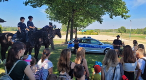 grupa dzieci, w tle radiowóz policjant i 2 policjantów na koniach