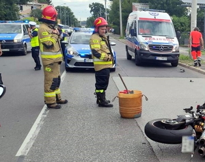 motocykl leży na jezdni, obok stoi policjant, który zapisuje dokumentację na protokole i strażacy, w tle radiowozy i karetka pogotowia