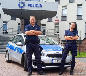 policjant i policjantka na tle Komendy Miejskiej Policji w Częstochowie pozują do zdjęcia stojąc przy radiowozie