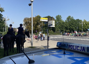 policjanci na koniach stoją za zaparkowanym radiowozem, patrzą w stronę stojących przed przejściem dla pieszych pielgrzymów