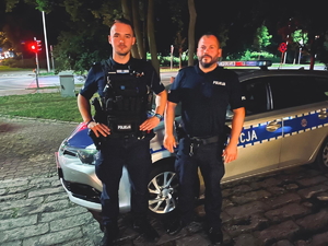 2 umundurowanych policjantów pozuje do zdjęcia na tle radiowozu