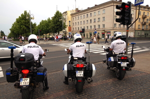 3 policjantów na motocyklach stoi przed skrzyżowaniem - ustawieni są tyłem do fotografującego, patrzą w kierunku przejścia dla pieszych