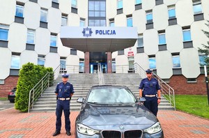 2 umundurowanych policjantów stoją po lewej i prawej stronie nieoznakowanego radiowozu, pozując na tle częstochowskiej komendy