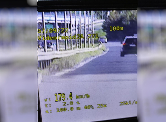zdjęcie z policyjnego wideorejestratora, na zdjęciu w tle pojazd koloru ciemnego, na pierwszym planie w dolnym roku wyświetlona jest prędkość kontrolowanego pojazdu i przedstawia zapis 179,4 km/h