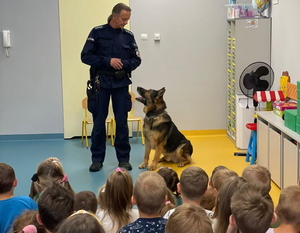 policjant z psem służbowym rozmawia z dziećmi, które siedzą przed nim na dywanie