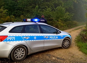 radiowóz zaparkowany na obszarze leśnym, przed nim stoi umundurowany policjant