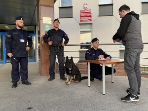 2 policjantów i pies służbowy podczas sprawdzania wchodzących na teren komendy rezerwistów stoją na schodach przed głównym wejściem