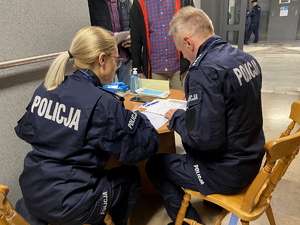 policjant i policjantka siedzą przy stoliku w holu komendy podczas sprawdzania wchodzących na teren komendy rezerwistów