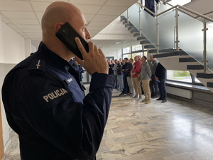 policjant na pierwszym planie rozmawia przez telefon, w tle stoją rezerwiści na korytarzu komendy