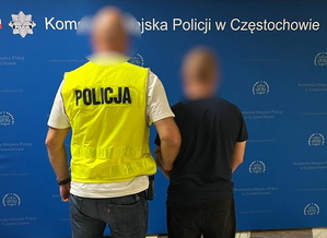 policjant w kamizelce odblaskowej trzyma pod rękę zatrzymanego w budynku komendy