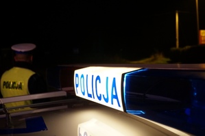 zdjęcie zrobione w porze wieczorowej - widać podświetlony napis policja na radiowozie, w tle policjant w kamizelce odblaskowej