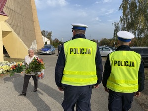 2 policjantów w kamizelkach odblaskowych i białych czapkach patrolują pieszo rejon cmentarza