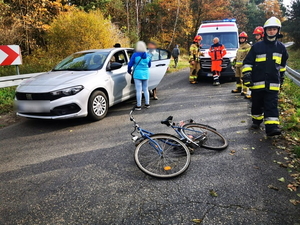 miejsce kolizji - srebrny fiat stoi na drodze, obok kobieta, na jezdni leży rower, w tle widać ratownika medycznego i straż pożarną