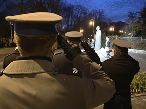 na pierwszym planie zbliżenie na salutującego policjanta, w tle pomnik księdza Jerzego Popiełuszki