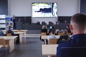 uczniowie siedzą w ławkach auli Komendy Miejskiej Policji w Częstochowie, patrzą na ekran i na podest, gdzie siedzą zaproszeni goście