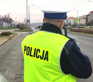 na zdjęciu policjant z ruchu drogowego ubrany w kamizelkę odblaskową koloru żółtego trzyma w ręce radar i mierzy prędkość pojazdom w rejonie przejścia dla pieszych, w tle widać radiowóz