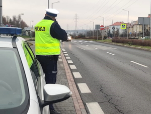 na zdjęciu policjant z ruchu drogowego ubrany w kamizelkę odblaskową koloru żółtego trzyma w ręce radar i mierzy prędkość pojazdom w rejonie przejścia dla pieszych, w tle widać radiowóz