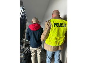 Na zdjęciu widać policjanta stojącego plecami w odblaskowej kamizelce, trzymającego za rękę mężczyznę również stojącego plecami z kajdankami na rękach założnych z tyłu.