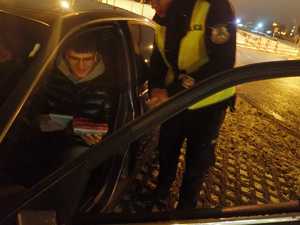 Policjant ruchu drogowego w umundurowaniu kontrolujący pojazd i jego kierowcę, kierowca trzyma w ręku apteczek oraz notatnik otrzymane od funkcjonariusza